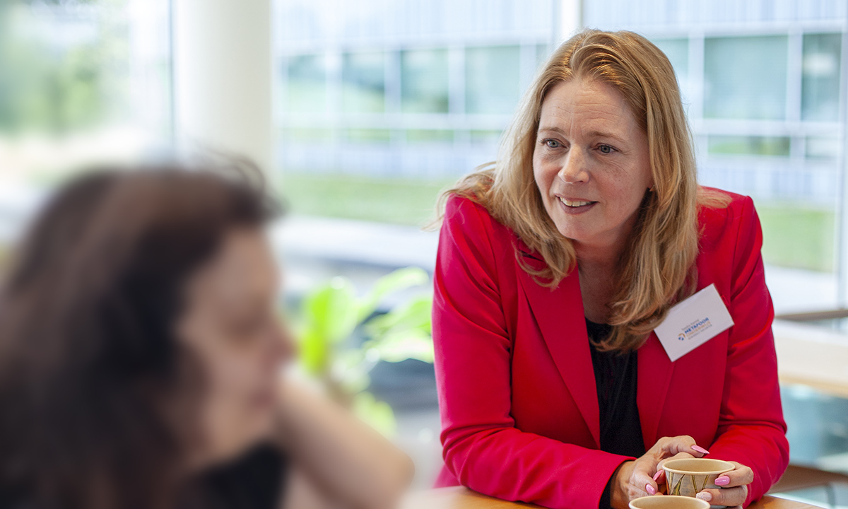 Sandra Samlal van Metafoor Onderwijs, gekleed in een roze colbert met een naambadge op, in een geanimeerd gesprek met een beker koffie in haar hand.