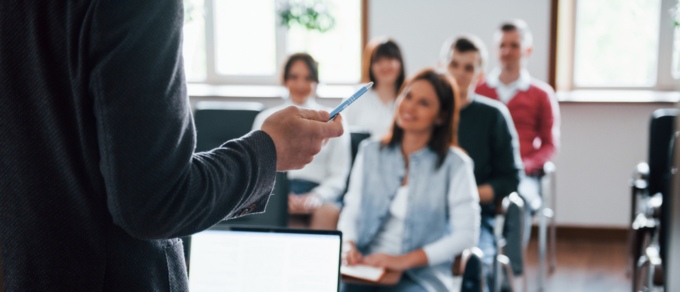 Docent met een pen in de hand voor een klas met aandachtig luisterende studenten, ter illustratie van praktische klassenmanagement tips.